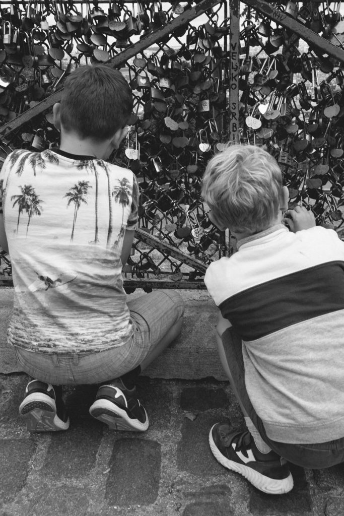 Deux enfants accroupis devant une grille couverte de cadenas, en noir et blanc