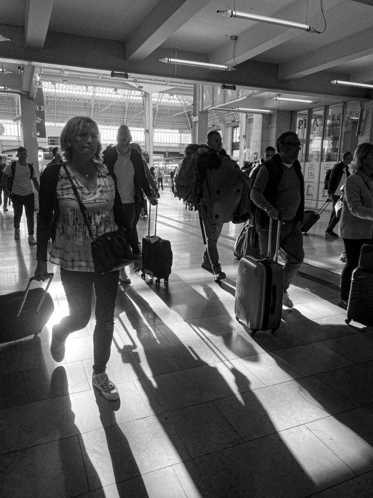 Des voyageurs traversent une gare à contre-jour, en noir et blanc